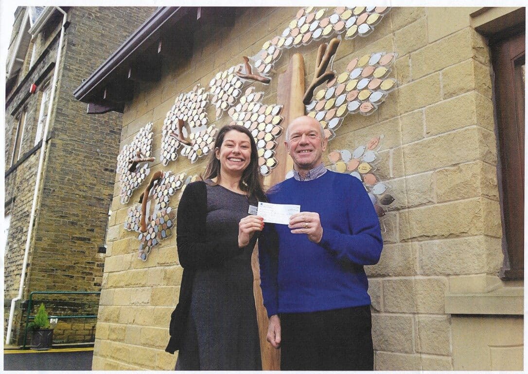 Colour photo of a man and a woman stood smiling outside a building, holding a cheque