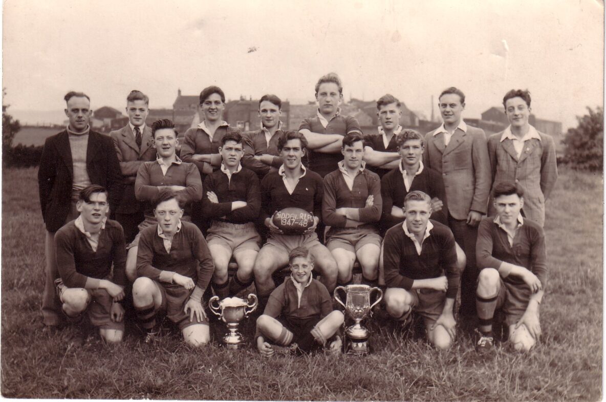 sepia team photo with three rows of players in their kits and two trophies on the ground in front