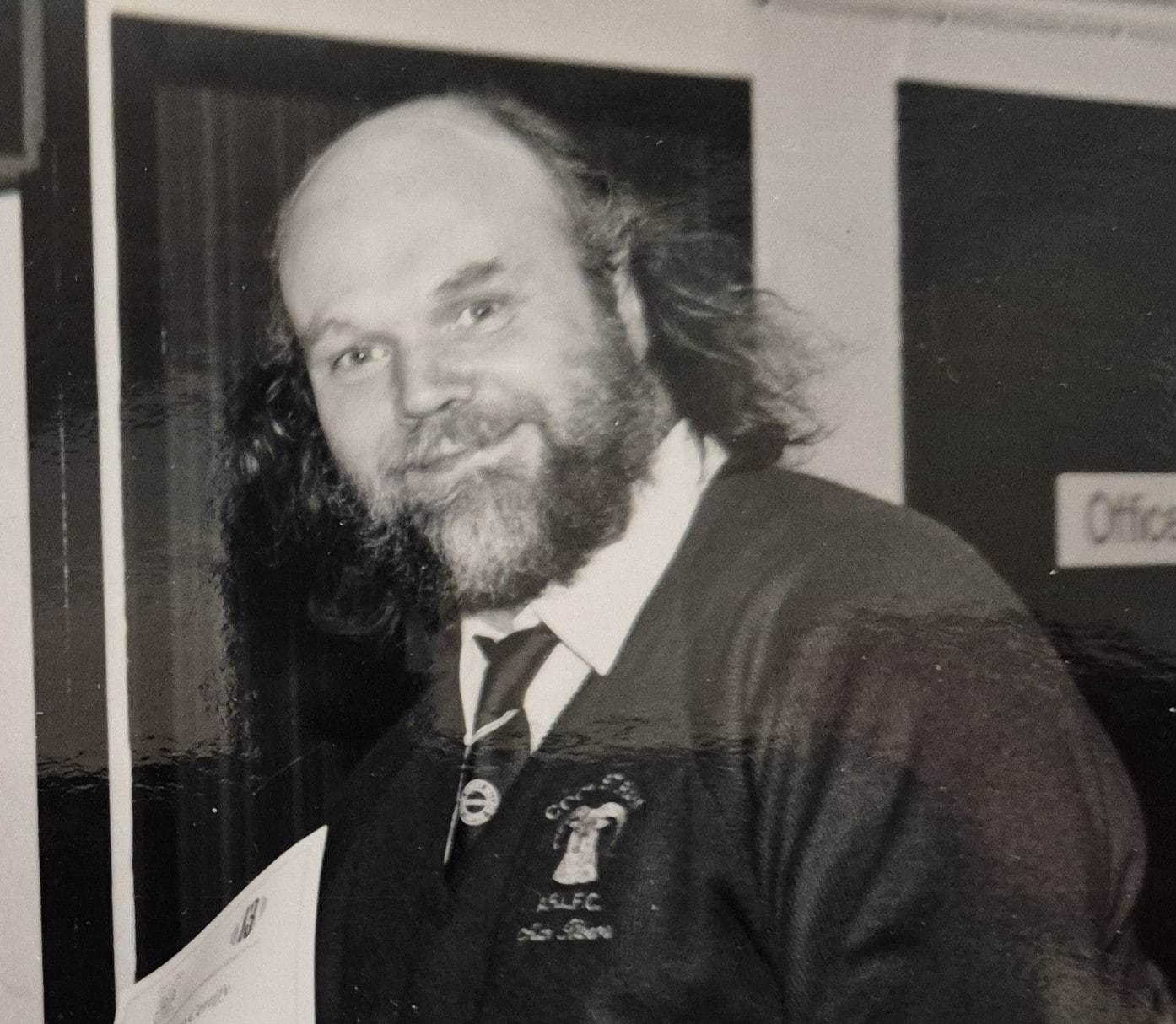 black and white photo of Alan Rivers who is smiling, has a beard and is wearing a light shirt, tie and cardigan
