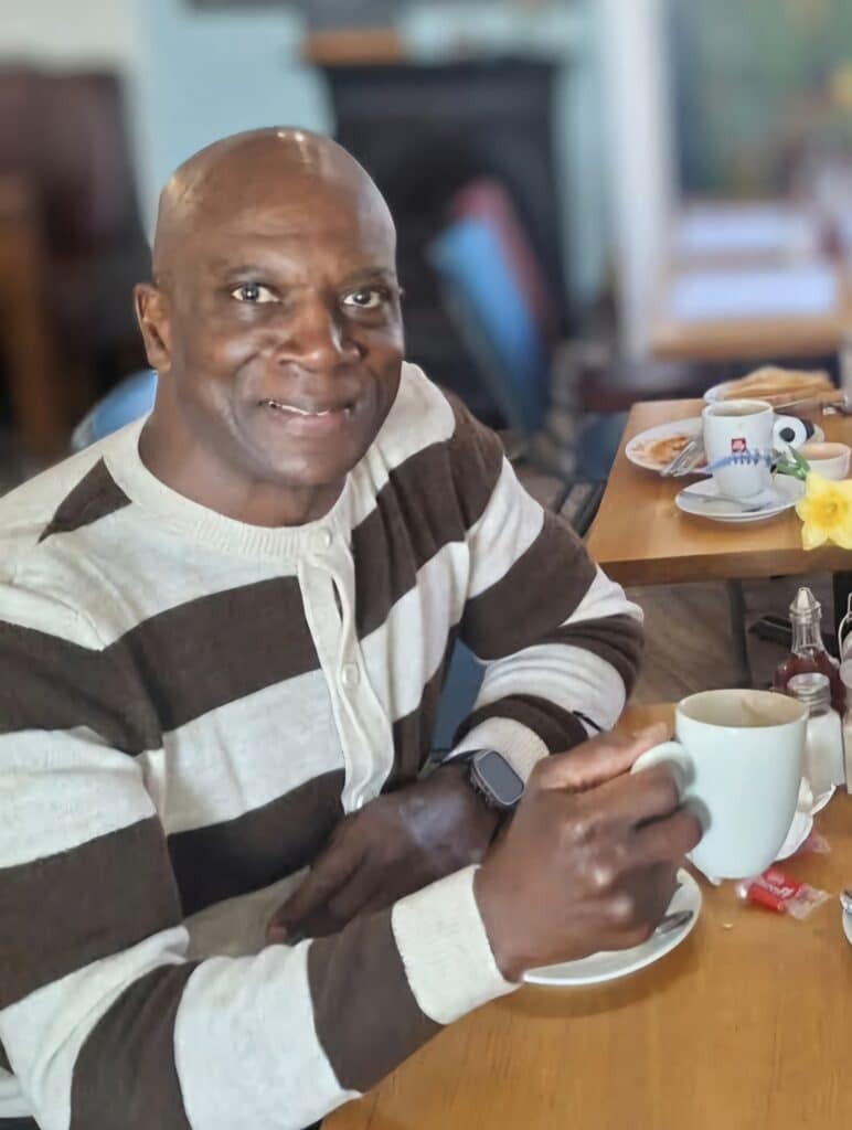 colour photo of Wilf George, who is smiling and holding a mug, wearing a cream and brown stripey long sleeved top