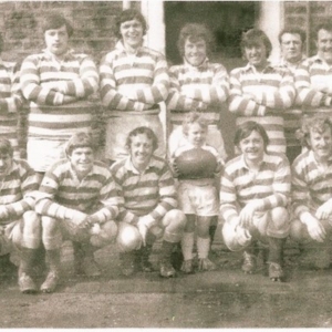 old photo of a team in two rows, wearing stripey kits, standing in front of a building. A small child holds the ball in the middle of the photo
