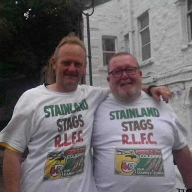tow people wearing white Stainland Staf R.L.FC t-shirts, they are smiling, the person on the left has their arm over the person on the right-who is also wearing glasses.