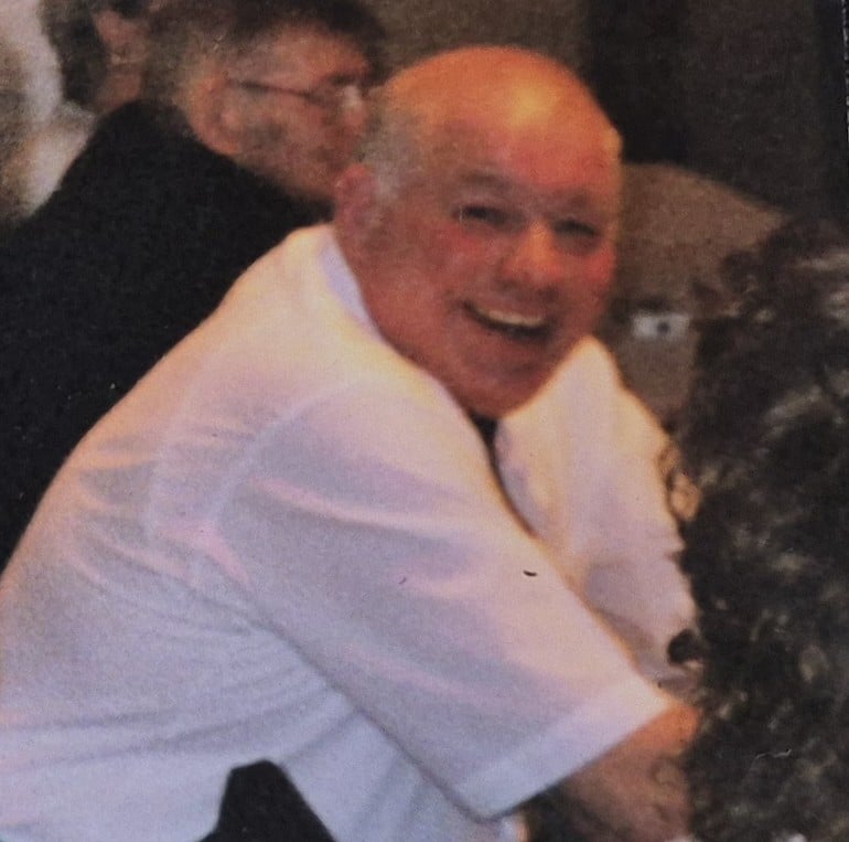 colour photo of John Sutcliffe who is smiling and wearing a short-sleeved white shirt, looking over their right shoulder to the camera