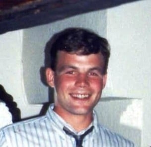 colour photo of Adam Greenwood who is smiling for the photo, wearing a white and blue stripey shirt and a dark-coloured tie and has short brown hair