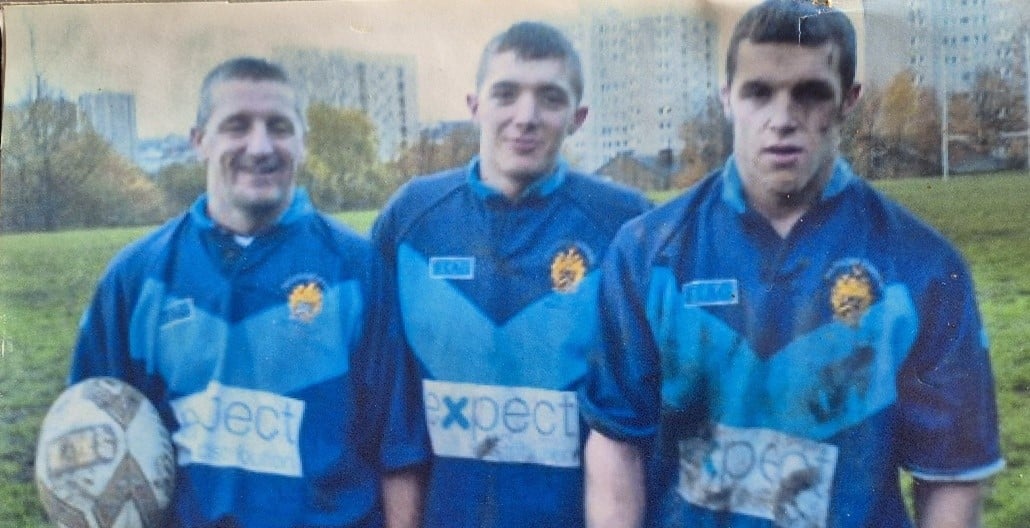 colour photo of Andrew with his sons Graham and Luke, wearing blue rugby kits on a pitch, Andrew is holding a rugby ball