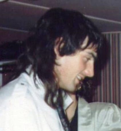 colour side profile photo of Gary Richardson who has long brown hair in a sort of mullet-style and is smiling, wearing a white top 
