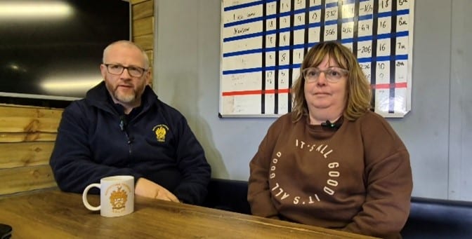 colour photo of Paul and Samantha Farrell, who are sat at a table, Paul is wearing glasses and a dark-coloured jacket, Samantha is wearing glasses and a brown sweater 