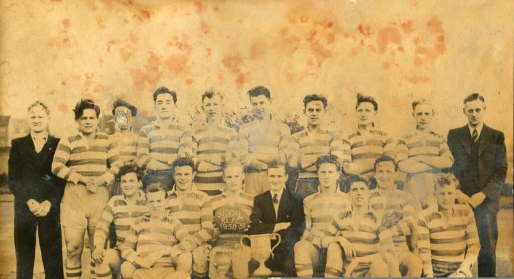 sepia photo of Siddal team in 1951, platers are in two rows, wearing stripey kits and there is a person either side wearing a dark suit, and one sat in the middle holding the trophy cup