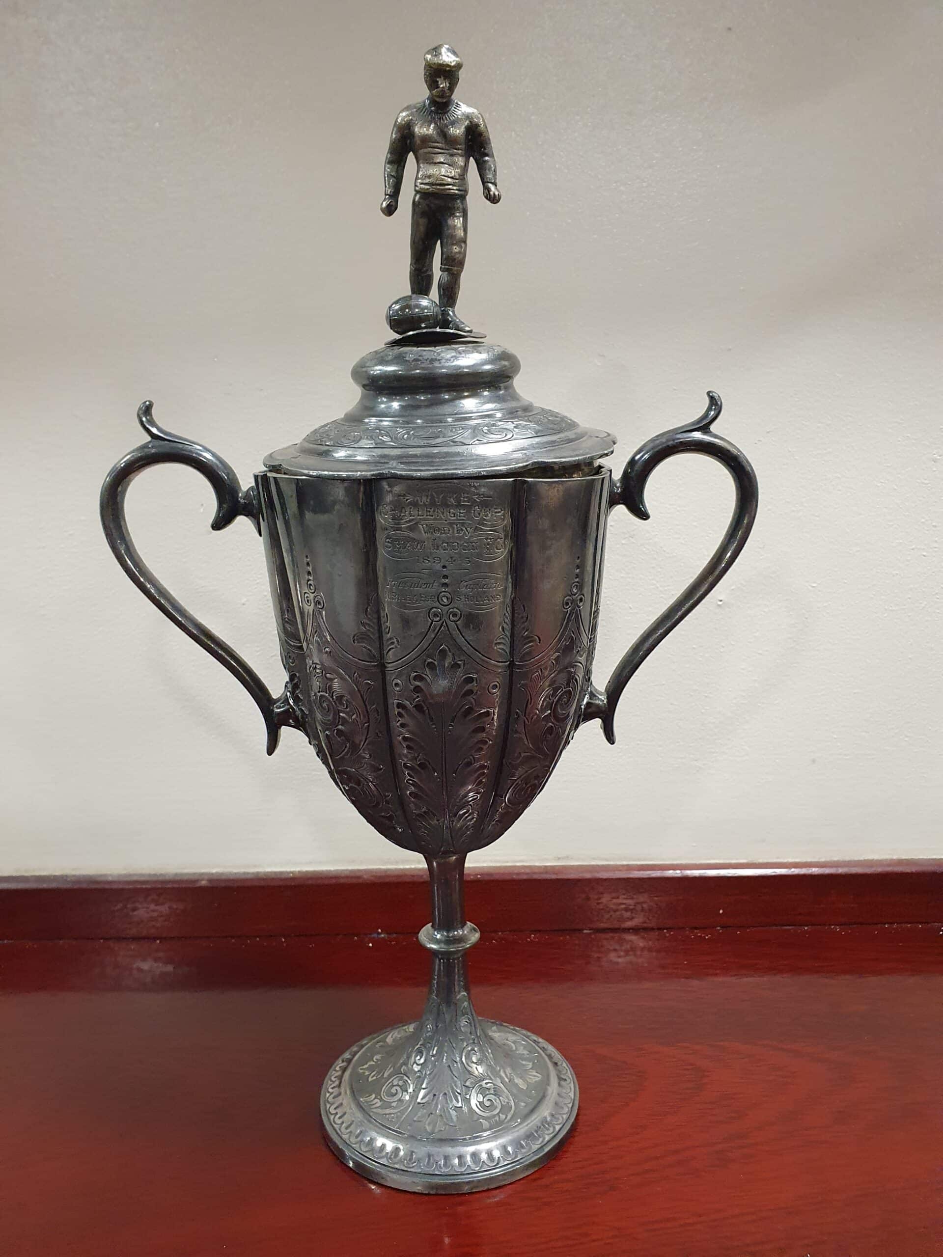 colour photo of Wyke Challenge Cup- a silver metal cup trophy with a player figure on top, against a white wall, sitting atop a varnished wood surface