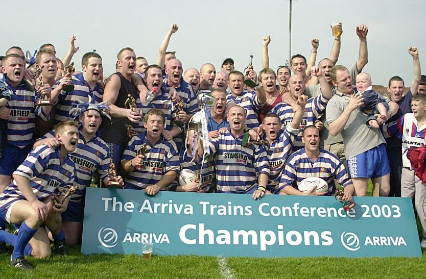 colour photo of the Siddal team celebrating on the pitch in a group, in front of the Arriva Trains Conference Champions banner