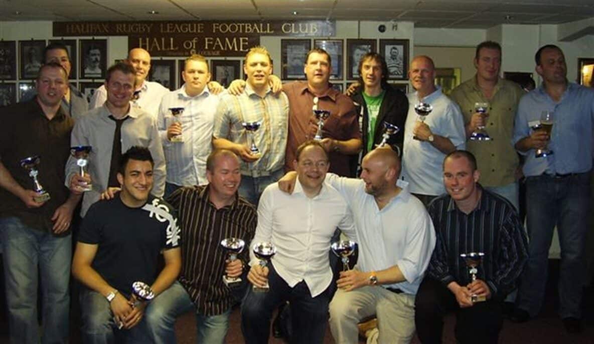 colour photo of a group of people wearing shirts, holding small trophy cups, standing in two rows inside a building
