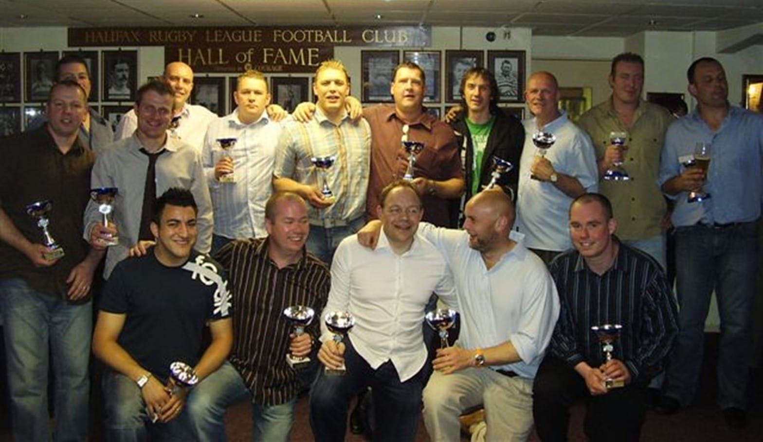 colour photo of a group of people wearing shirts, holding small trophy cups, standing in two rows inside a building 