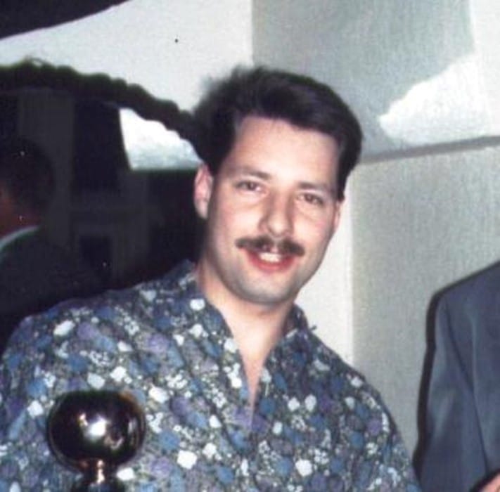 colour photo of Paul Glover, who is smiling wearing a blue shirt with a white pattern on it, holding a trophy, they have a moustache and short brown hair 