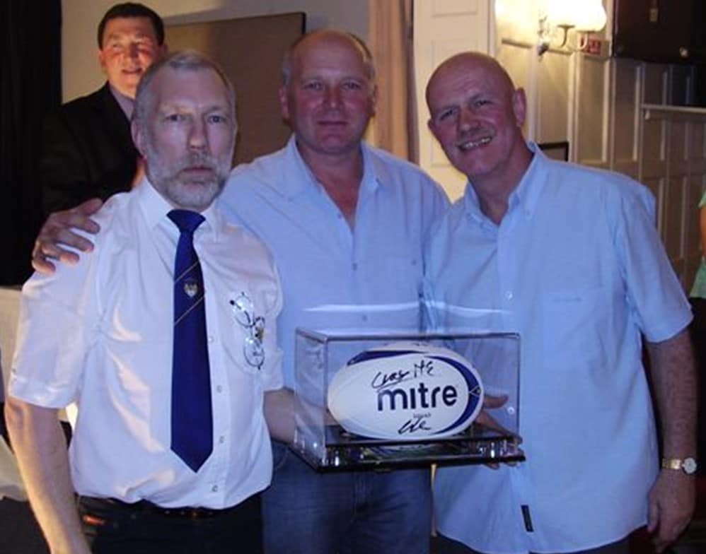 colour photo of three people wearing shirts, smiling for the camera, the middle person is wearing a mitre rugby ball in a glass box 