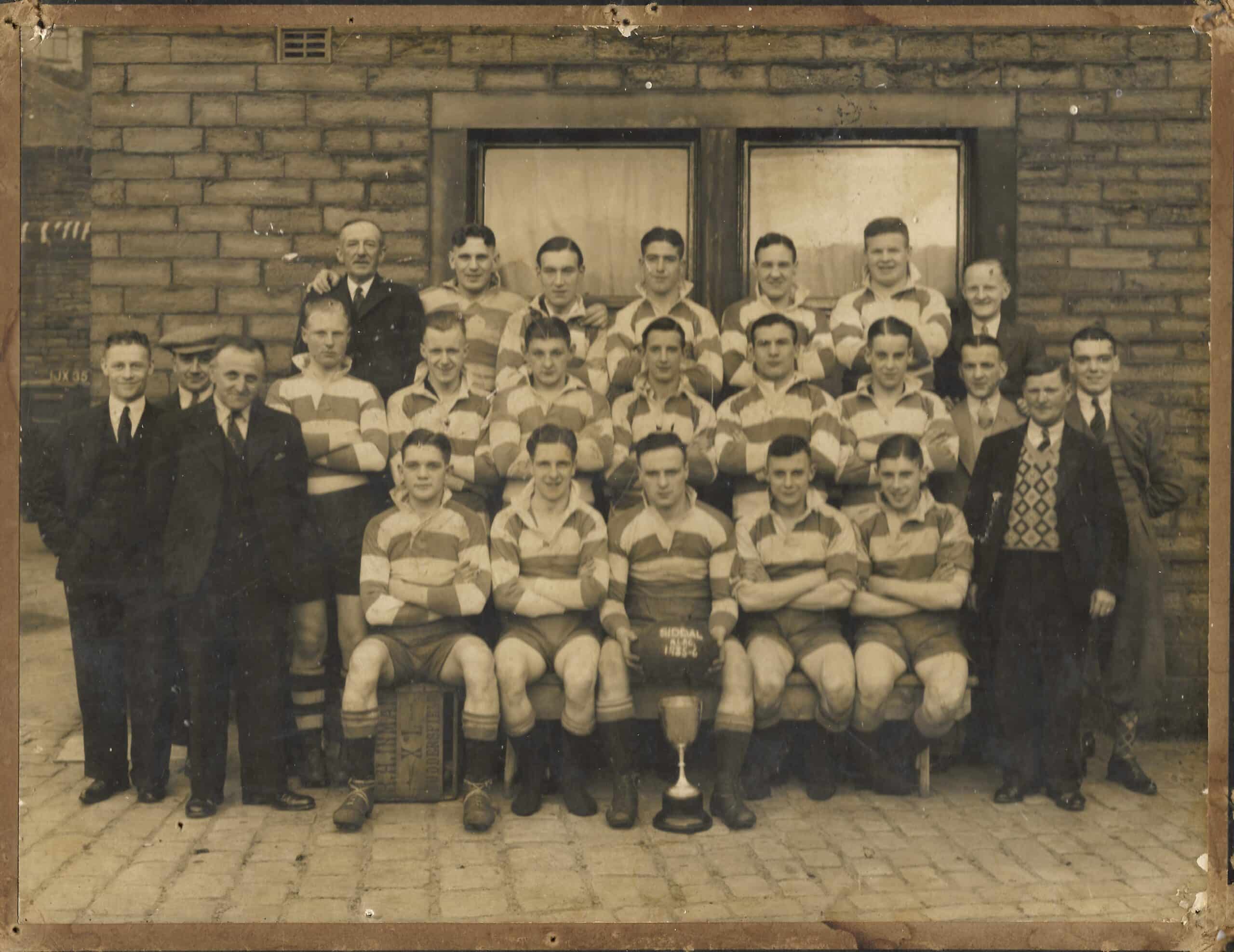 sepia scan of the Siddal 1935-5 team, the players are wearing stripey kits and in three rows in front of a building, with a few people either side, wearing dark suits