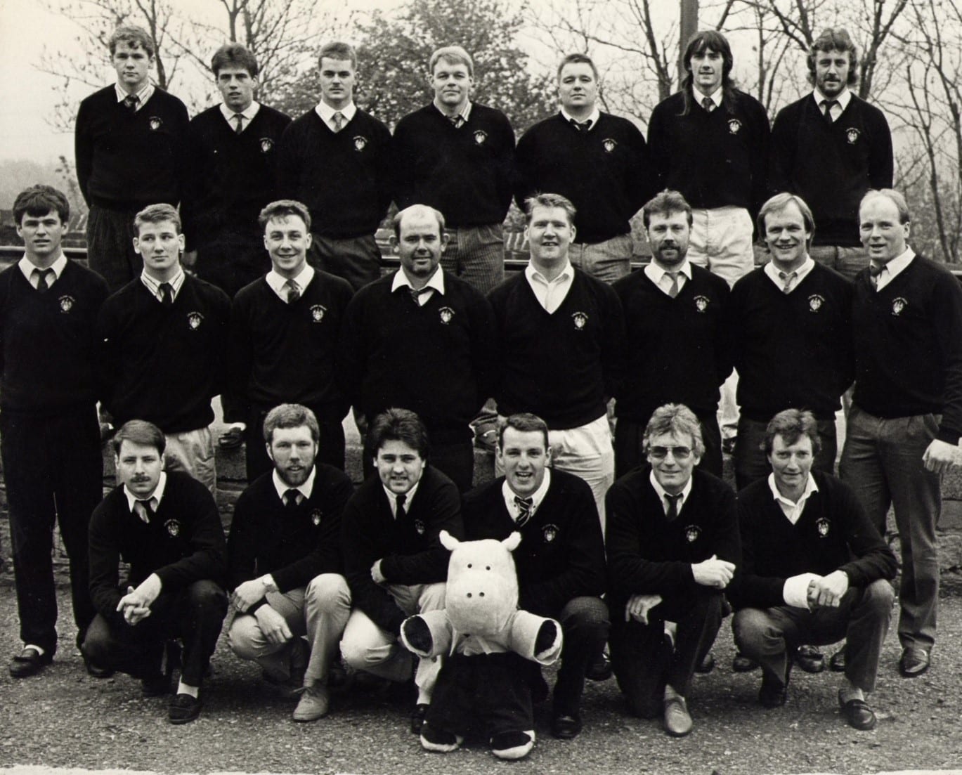 black and white photo of team wearing dark sweaters and ties and shirts, in three rows with a mascot plush at the front in the middle 