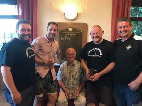 Two people stand either side of Barry, who is seated and they are all smiling. They are indoors at a club 