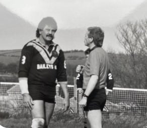 black and white photo of player Gary Ellis wearing their kit and talking to a referee. Gary has dark hair and a moustache