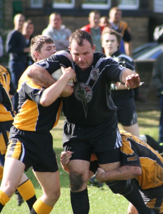 colour action shot photo of Greetland player Adam Greenwood, who is running with the ball and being tackled on either side 