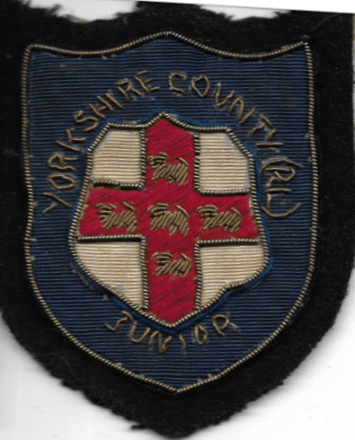 colour photo of an old Yorkshire County Junior sewn patch, which has a red cross in the middle and stitched lettering around it 